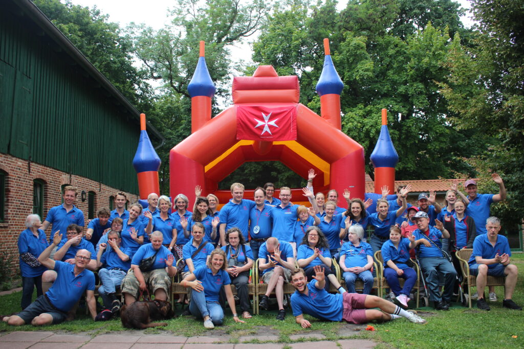 Gruppenbild der Integra-Freizeit: Gäste und Betreuende in blauen Shirts vor einer Hüpfburg mit Johanniterkreuz