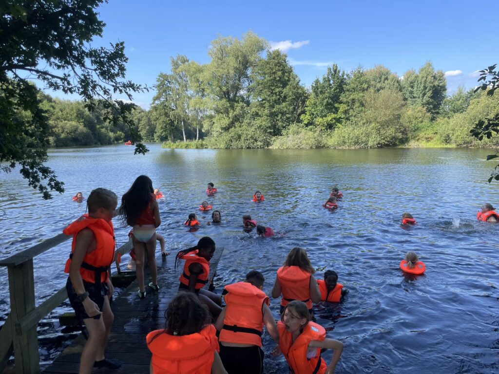 Kinder in orangen Schwimmwesten baden bei der Kinderfreizeit an einem See