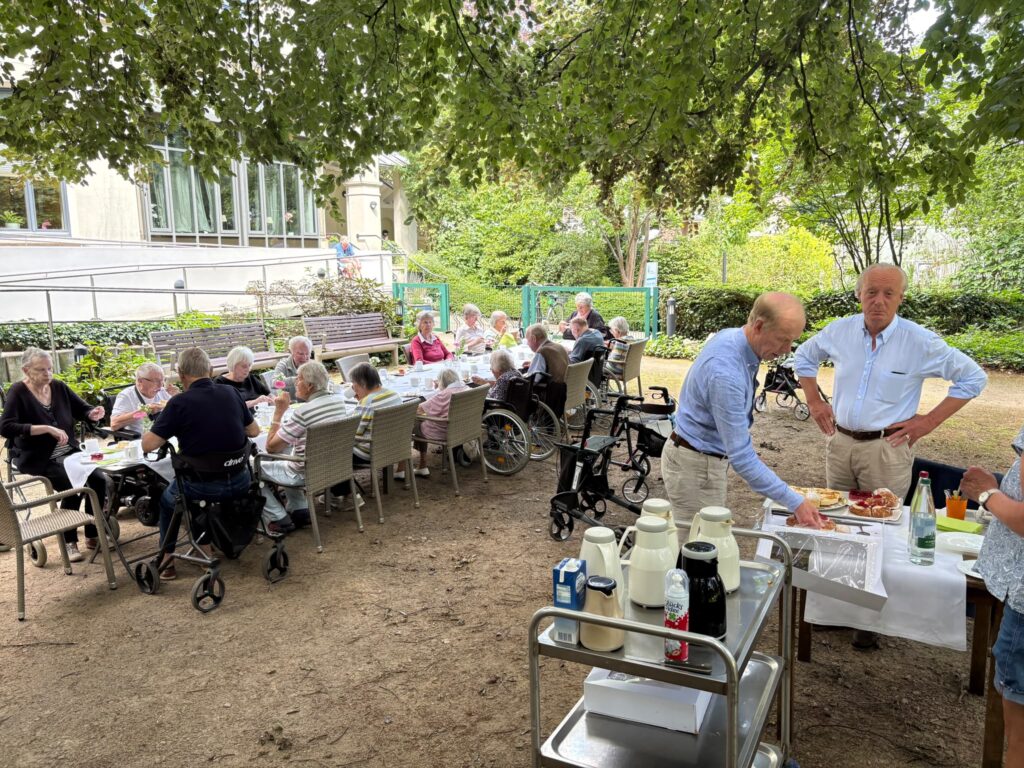 Gartenfest der Auguste-Viktoria-Stiftung: Bewohnerinnen und Bewohner an einer langen Kaffeetafel im Schatten